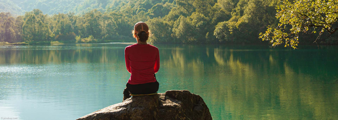 Person sitzt auf einem Stein und schaut über einen Bergsee - Symbolbild: Meditation