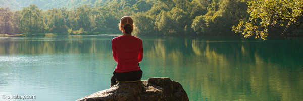 Person sitzt auf einem Stein und schaut über einen Bergsee - Symbolbild: Meditation